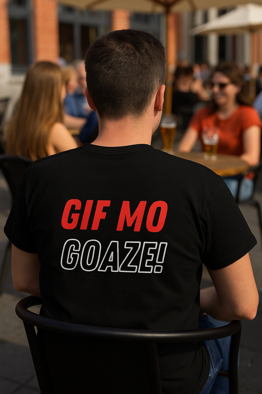 Persoon met zwart T-shirt met de tekst "GIF MO GOAZE!" zit op een zonovergoten terras in West-Vlaanderen, omringd door mensen die genieten van een drankje.

